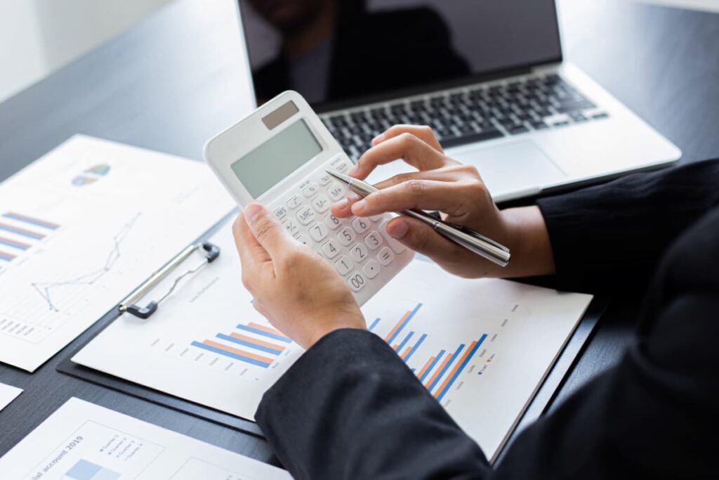 Pessoa calculando despesas fixas e variáveis com uma calculadora, gráficos financeiros e um laptop em uma mesa de escritório.