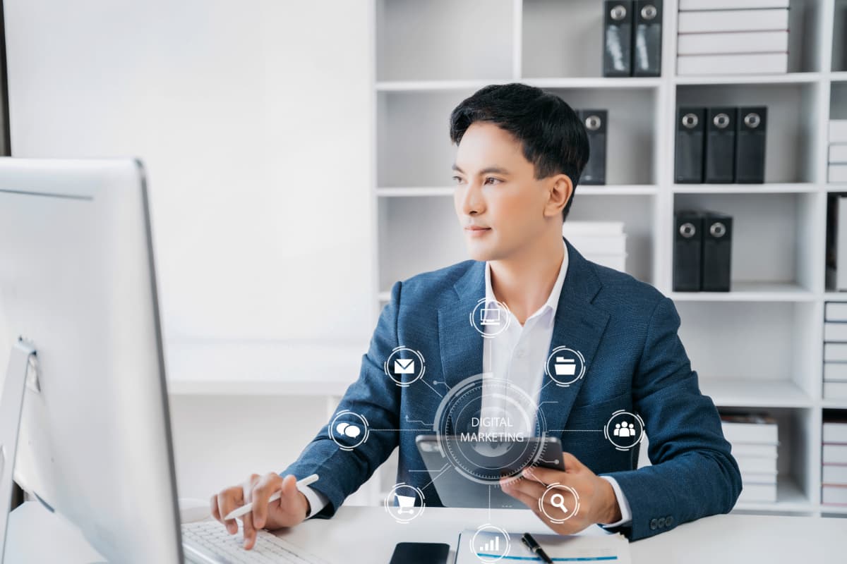 Profissional em terno trabalhando no laptop com ícones de marketing digital flutuando acima da mesa.