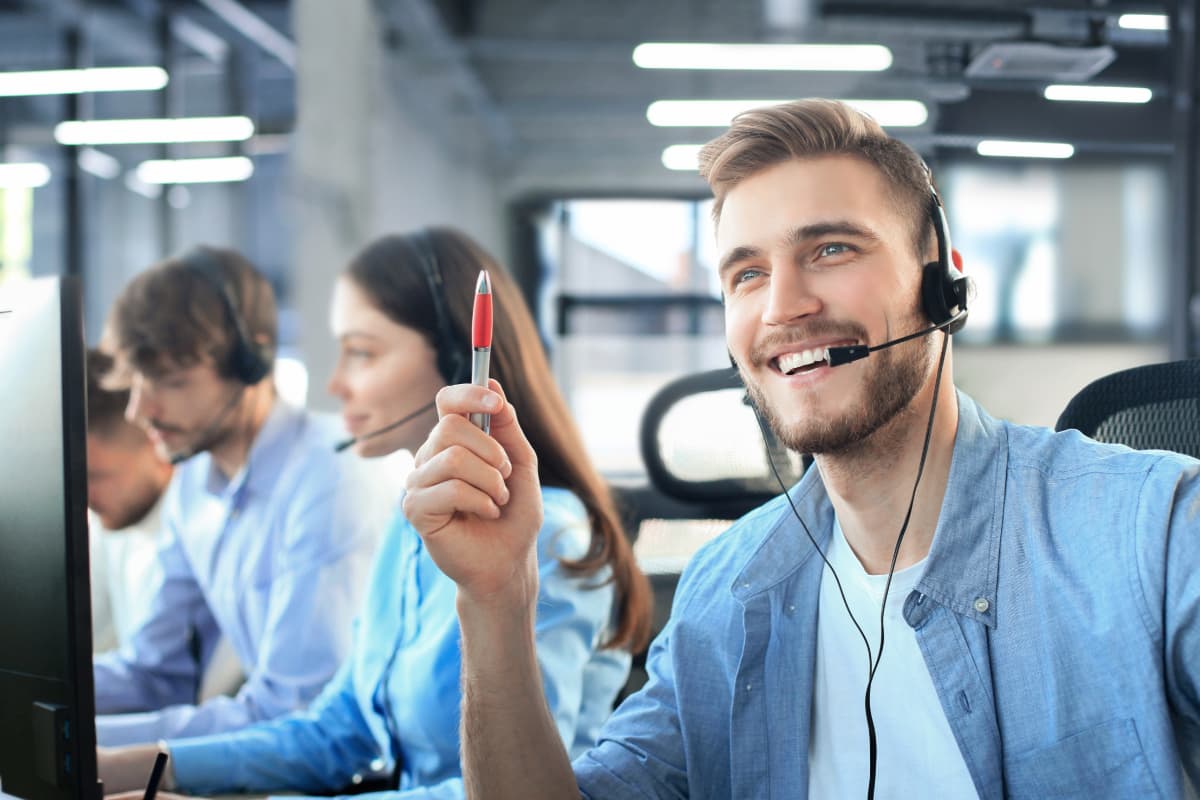 Enviado por Copilot:Pessoa com headset segurando uma caneta vermelha em um escritório, com colegas desfocados ao fundo.