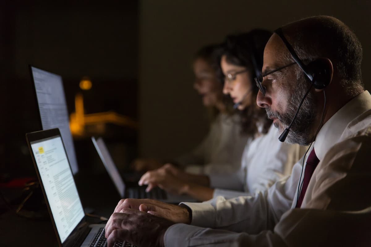 Profissionais em um call center usando tecnologia VoIP, com fones de ouvido e laptops, em um ambiente de baixa luz.