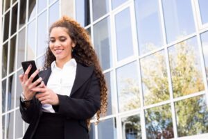 empresária de terno está na frente de um prédio olhando para o celular e busca entender sobre as politicas de uso de celulares corporativos