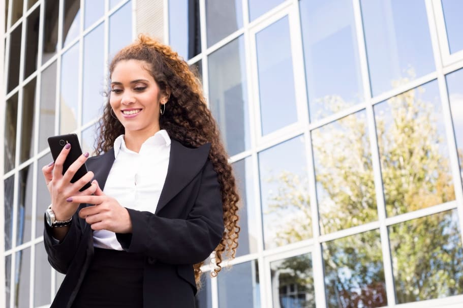 empresária de terno está na frente de um prédio olhando para o celular e busca entender sobre as politicas de uso de celulares corporativos
