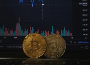 Fotografia de duas moedas de bitcoin em frente a uma tela com gráficos financeiros.