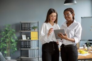 Duas mulheres sorrindo enquanto discutem estratégias de comunicação corporativa usando um tablet no escritório.