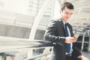 Homem de roupa social, usando um celular encostado em uma grade.