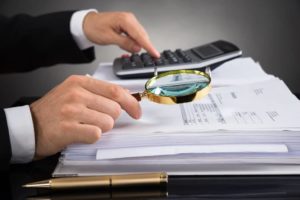 Homem de terno preto, segurando uma lupa com a mão direita e fazendo contas em uma calculadora com a mão esquerda.