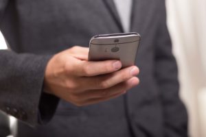 Homem de roupa social segurando um celular corporativo com a mão direita, e olhando para ele.